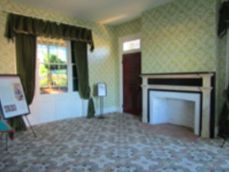 A room with pale green patterned wallpaper, a fireplace, dark green curtains over a window, and a patterned carpet.