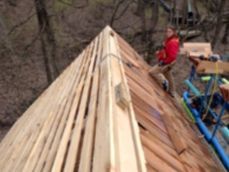A person in a red jacket installs cedar shake roofing on a sloped roof structure outdoors.