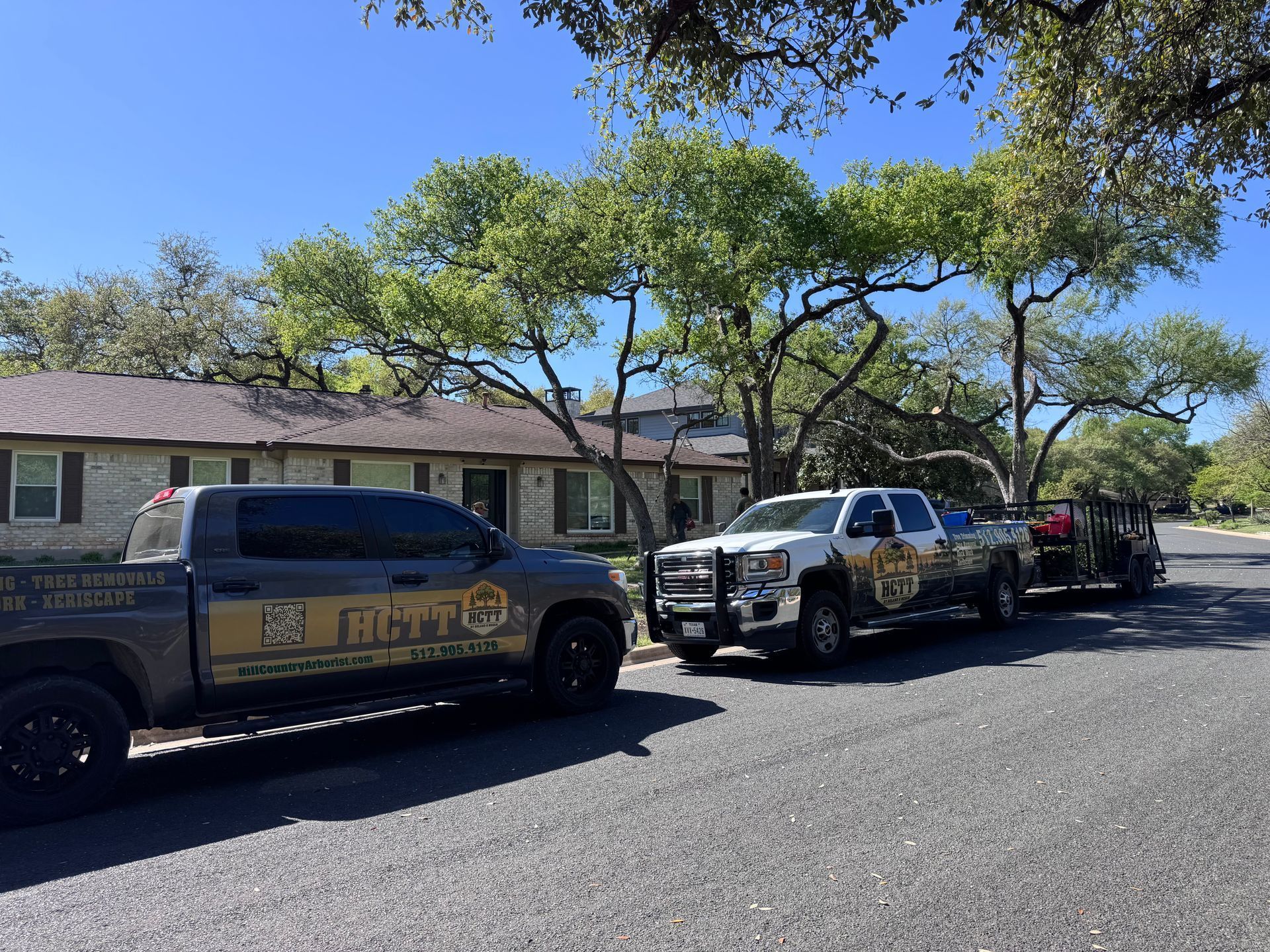 Two branded pickup trucks with trailers parked on a street in front of a stone house under a clear blue sky.