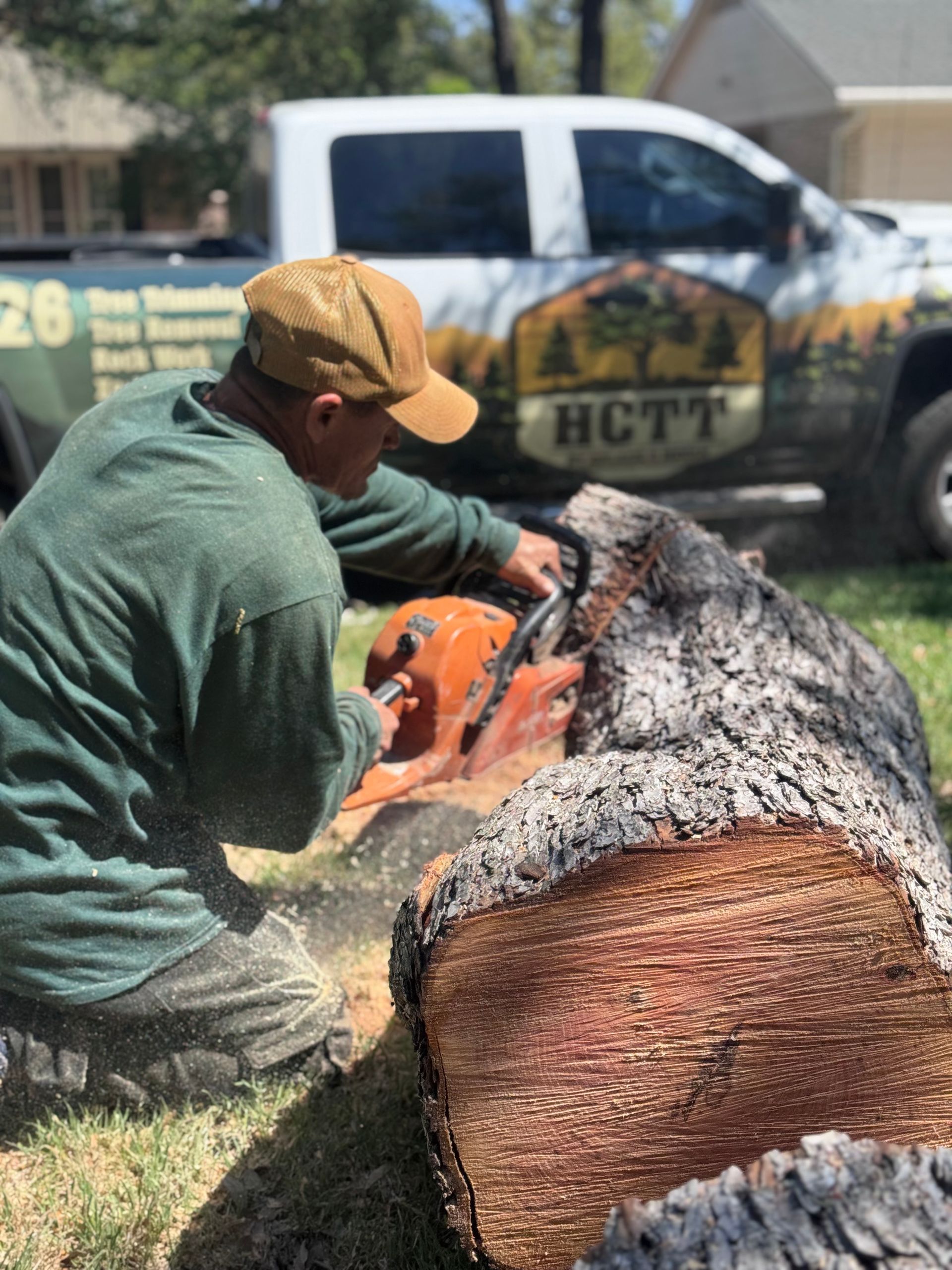 A person in a green shirt and tan cap kneels while using a chainsaw to cut a large log near a branded HCTT service truck.