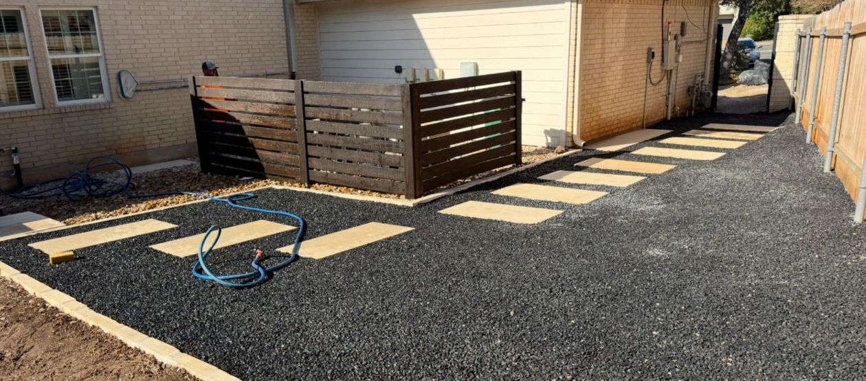 Backyard with black gravel, stepping stones, and a wooden fence. A blue hose lies on the ground.