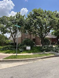 A brick house on a street corner with large trees, a street sign, and green bushes.