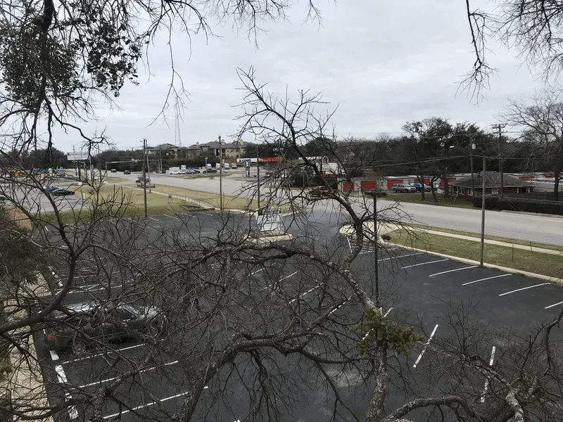 Overhead view of a parking lot and a street with businesses under an overcast sky, framed by bare tree branches.
