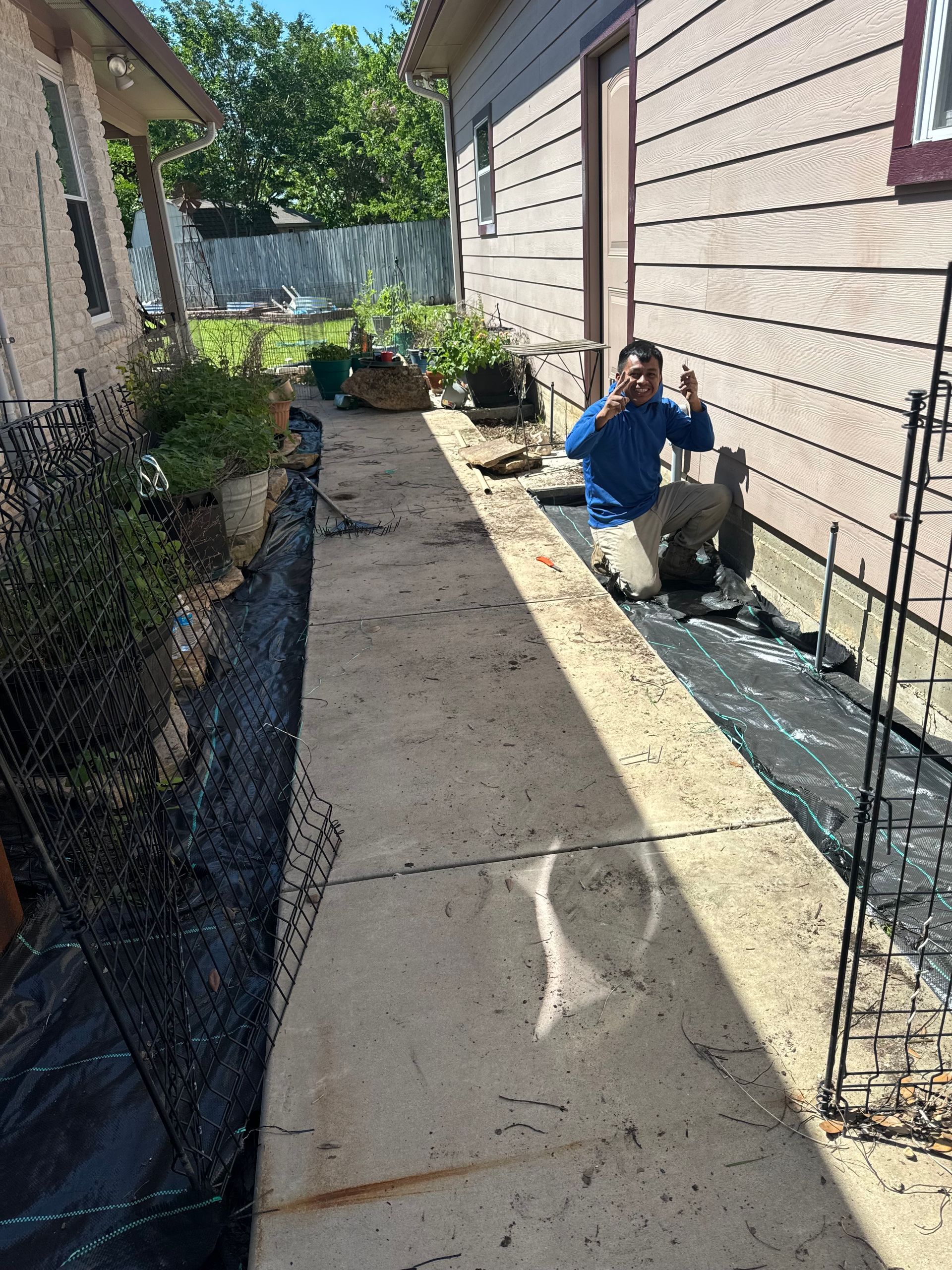 A person kneels near a building next to a walkway, holding up a thumbs-up. Black netting and landscape fabric are visible.