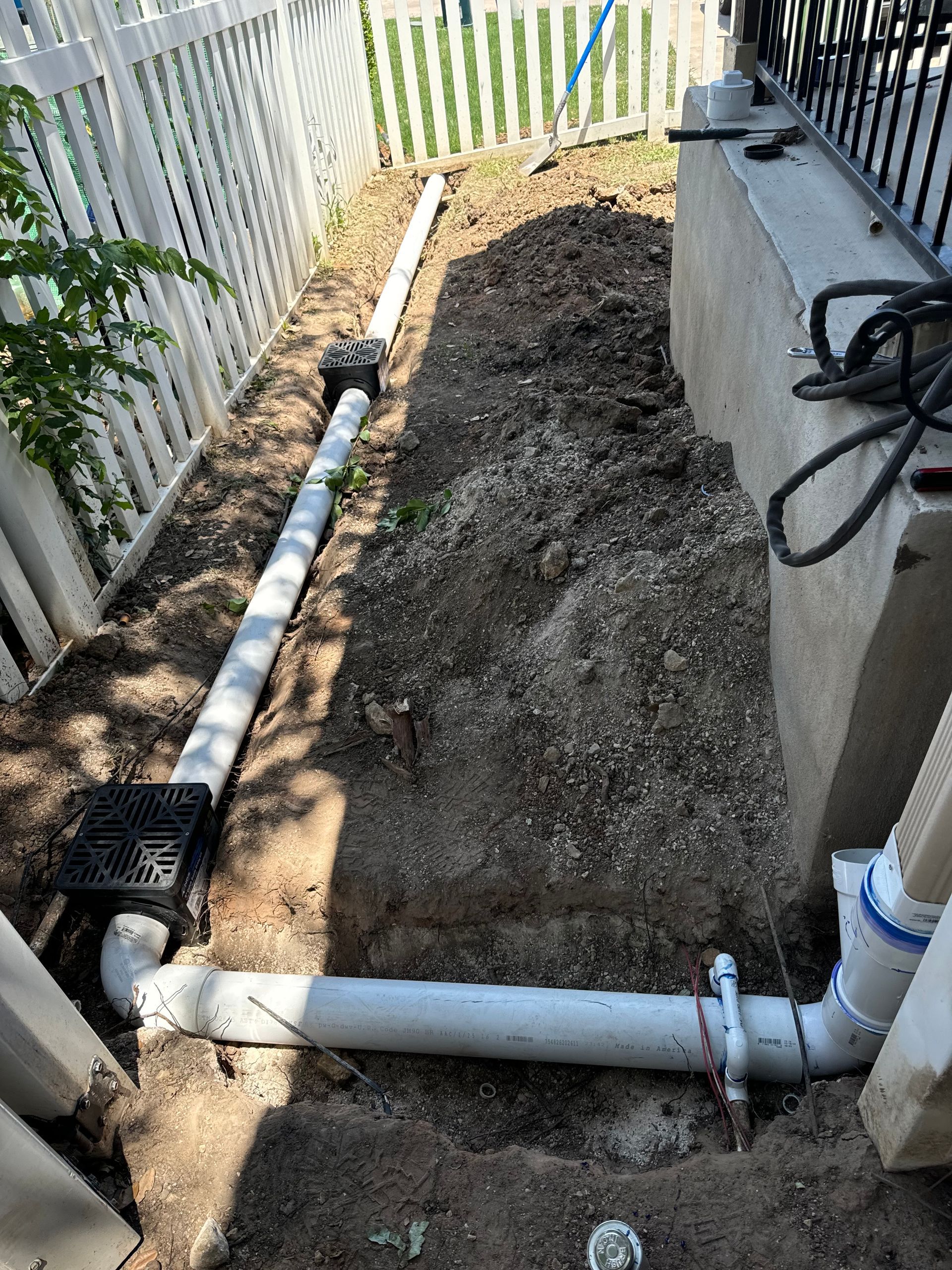 Drainage pipes being installed in a narrow dirt area next to a white fence and concrete foundation.