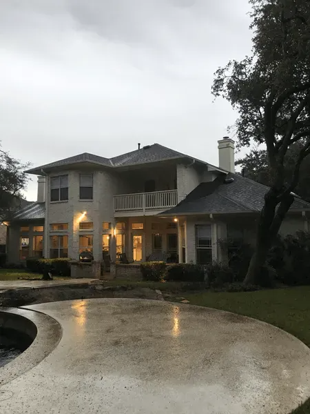 Two-story beige house with balcony, chimney, and wet patio under overcast sky.