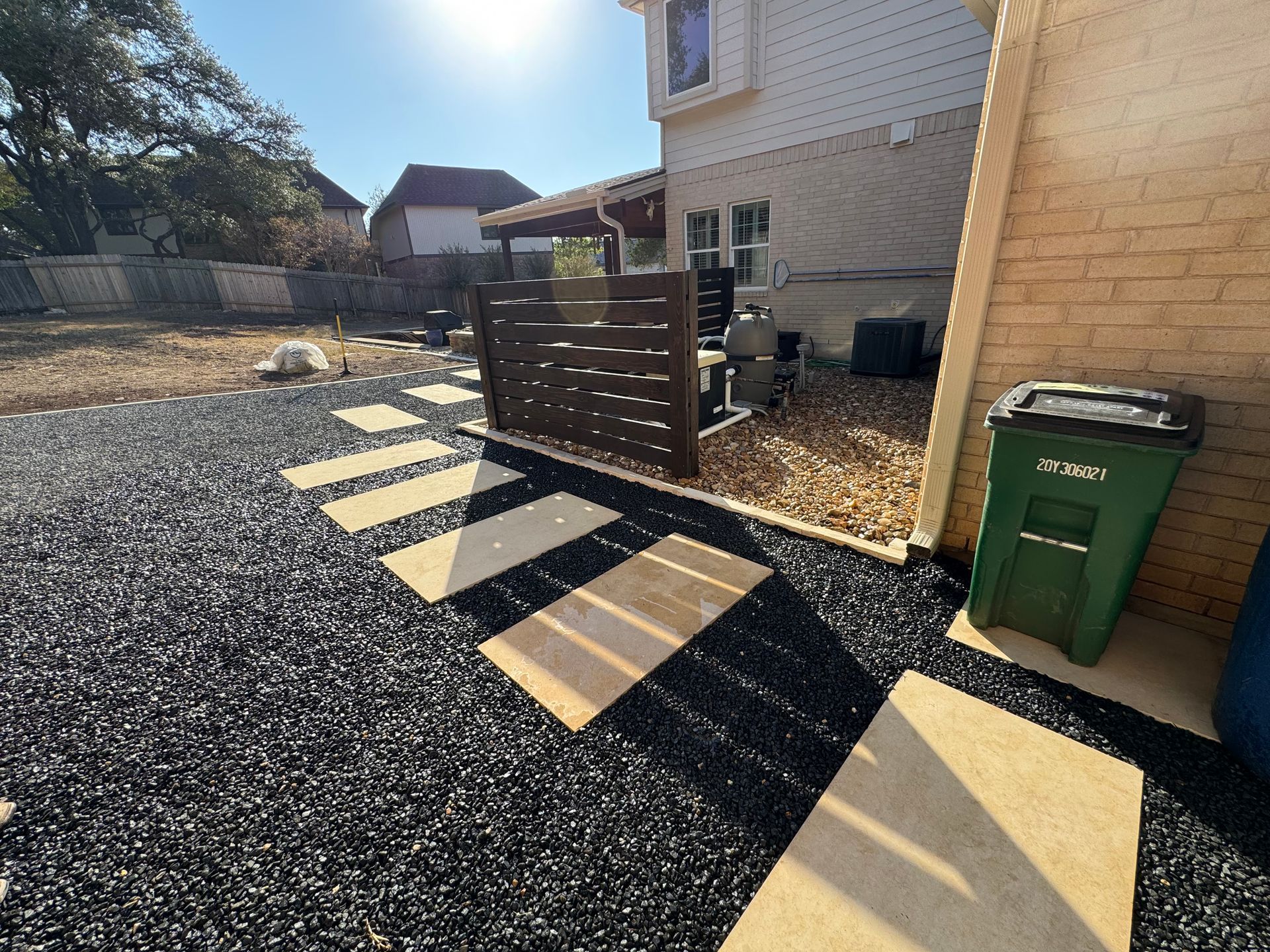 Backyard with stepping stones, black gravel, a wooden fence, and a green trash can.