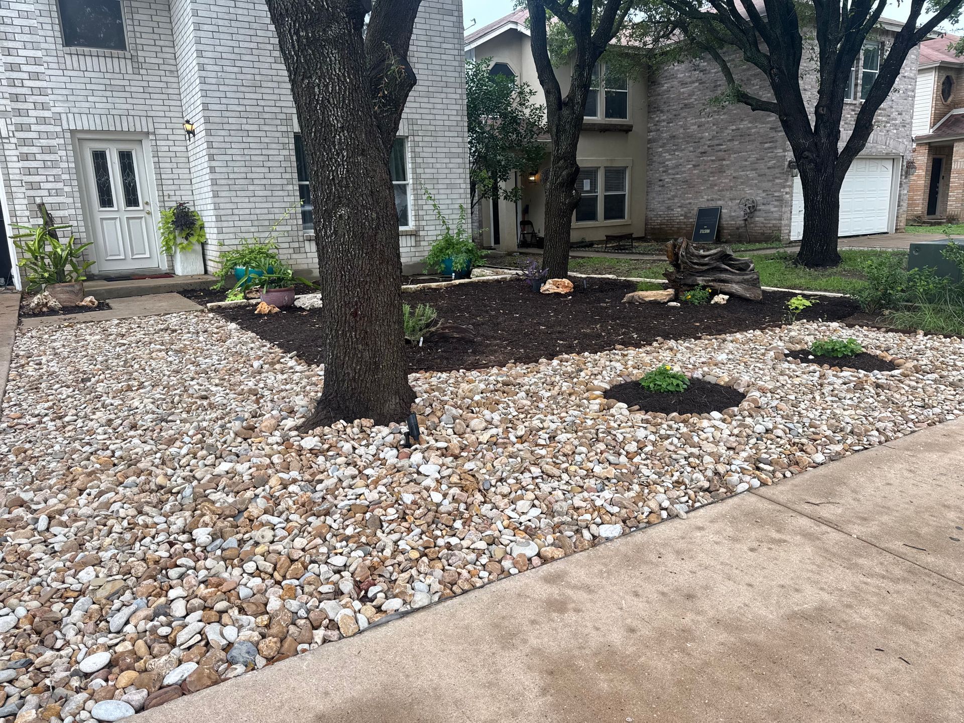 Stone-covered front yard with trees, plants, and a house with a white brick facade.