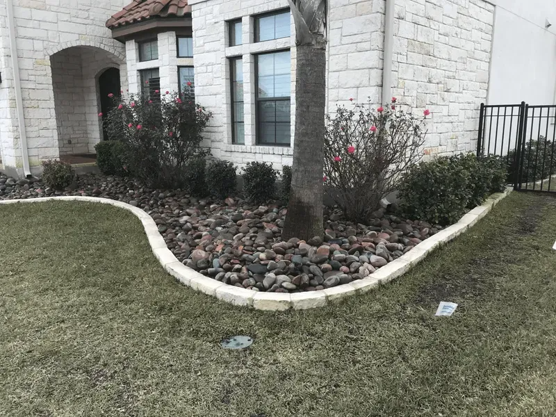 Stone house with landscaped yard, flower bed with rocks, bordered by light-colored stones, grass in foreground.