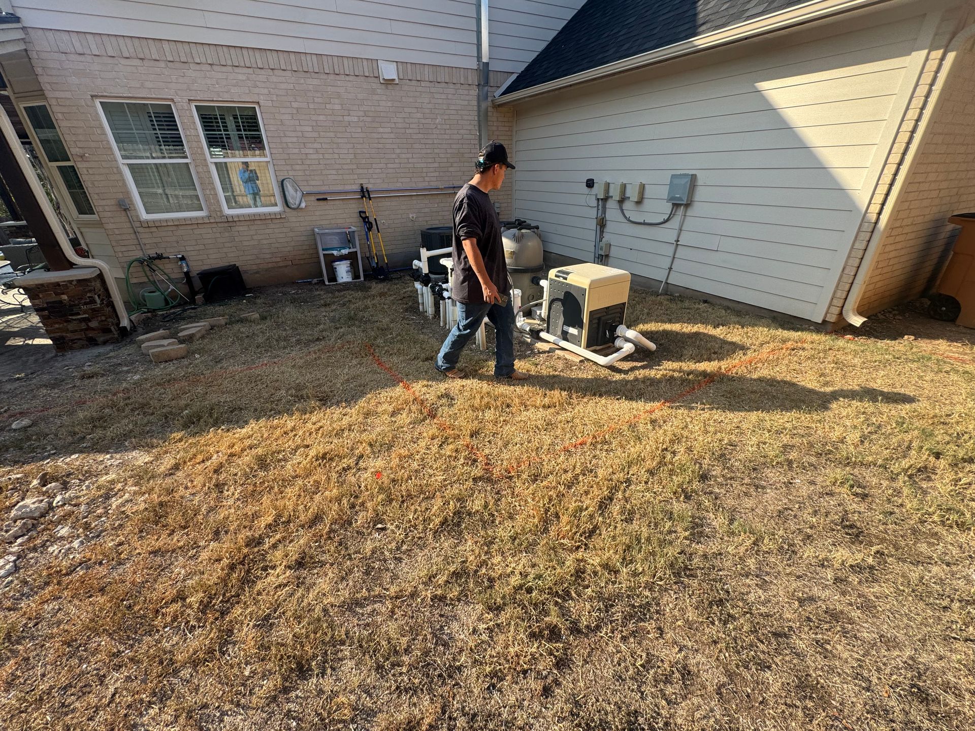 Man walking near pool equipment, next to a house with beige siding and windows. Dry grass in the yard.