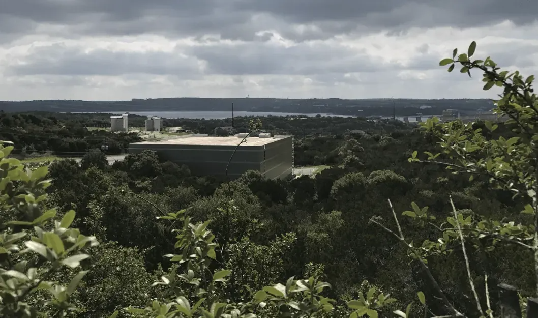 View from a high vantage point overlooking a large square building surrounded by trees, lake and cloudy sky.
