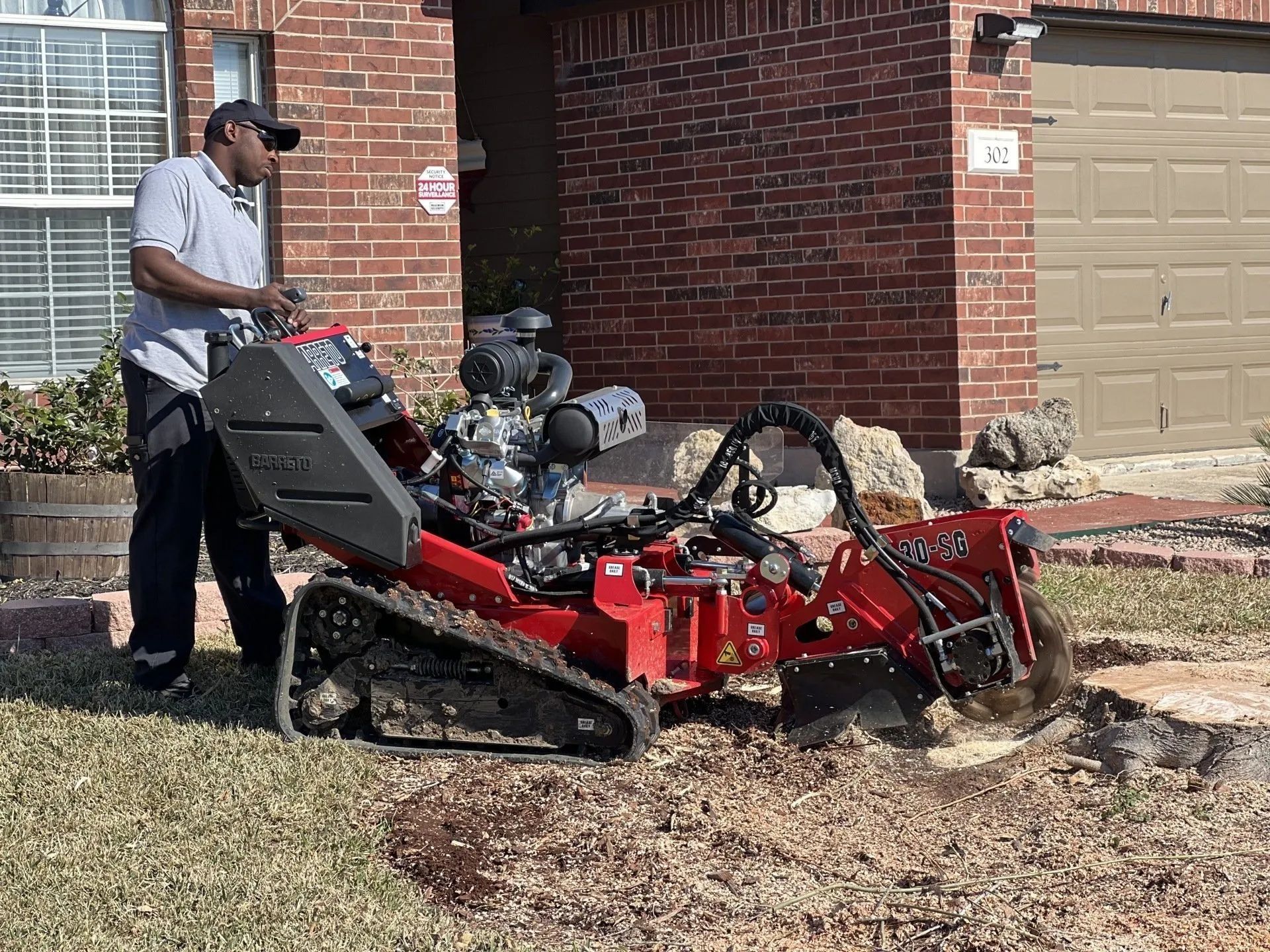 Man operating a red stump grinder on a residential lawn, brick house in background.