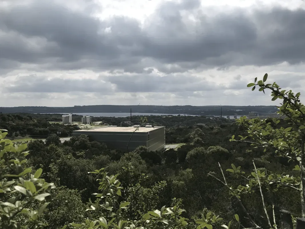 Overlook of a large, flat-roofed building and a distant body of water under a cloudy sky, with trees in the foreground.
