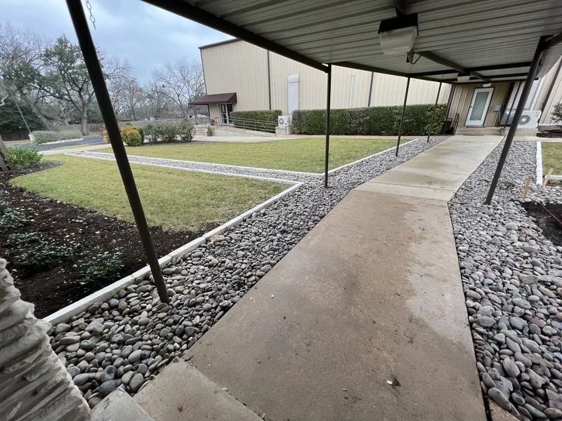 Concrete walkway under a covered area, bordered by gravel and grass, leading to a building entrance.