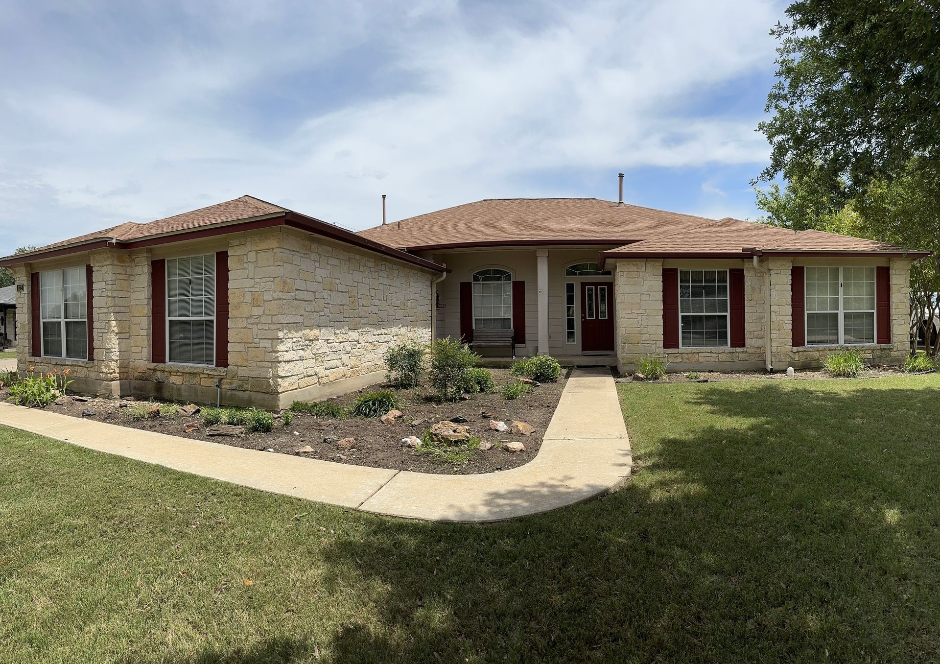 Beige brick house with red shutters and a brown roof under a cloudy sky.