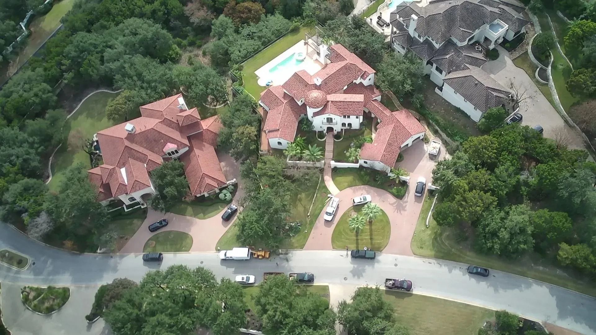 Aerial view of large houses with red tile roofs, a pool, and circular driveways surrounded by trees.