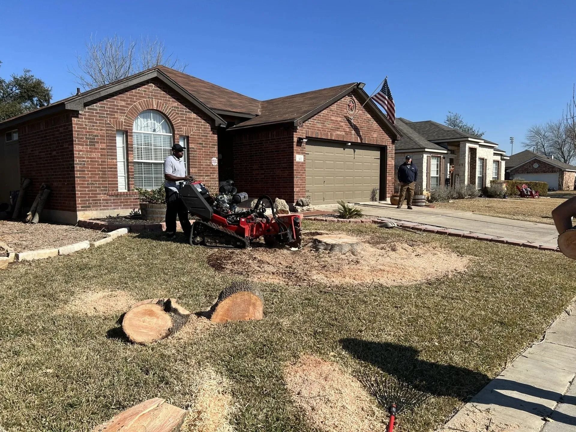 Man operating a stump grinder in front of a brick house. Wood shavings are scattered on the lawn.