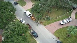 Aerial view: street with vehicles, including a truck, van, and bulldozer, near green trees and houses.