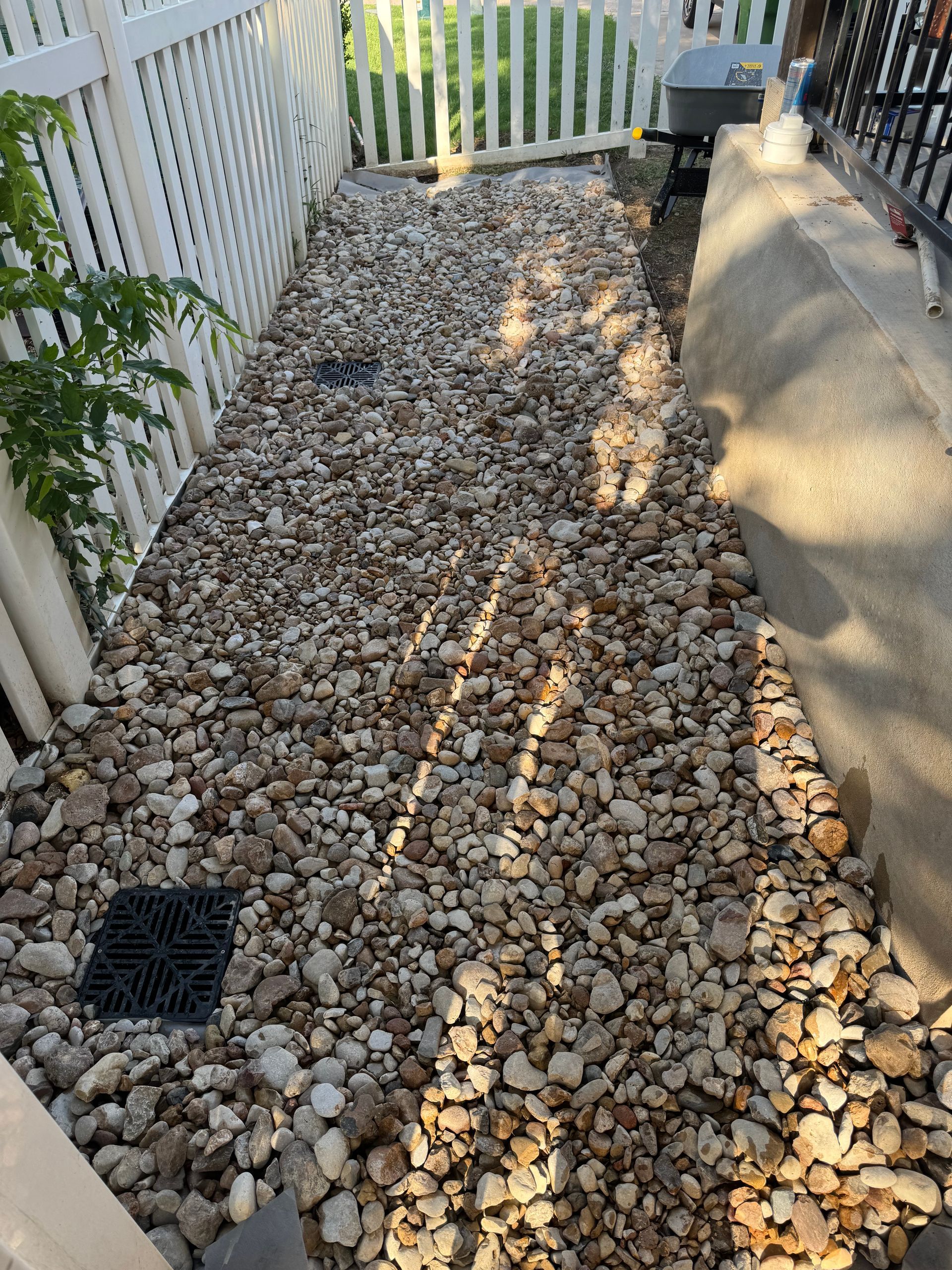 Gravel pathway between a white fence and concrete wall, leading towards a utility cart.