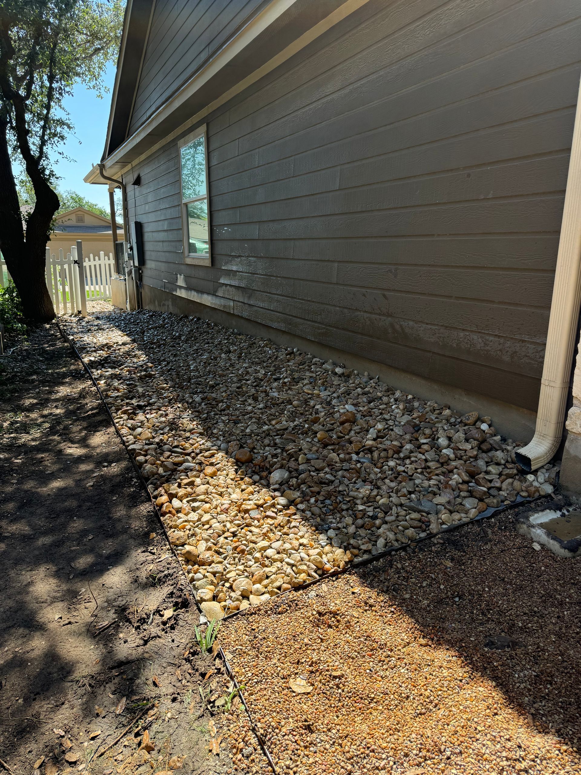 Side of a house with gravel bed. Brown siding, white trim, and a path of smaller gravel.