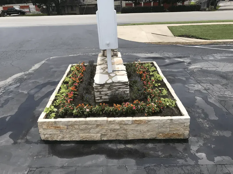 Stone planter box with colorful flowers at the base of a sign post, set on asphalt.