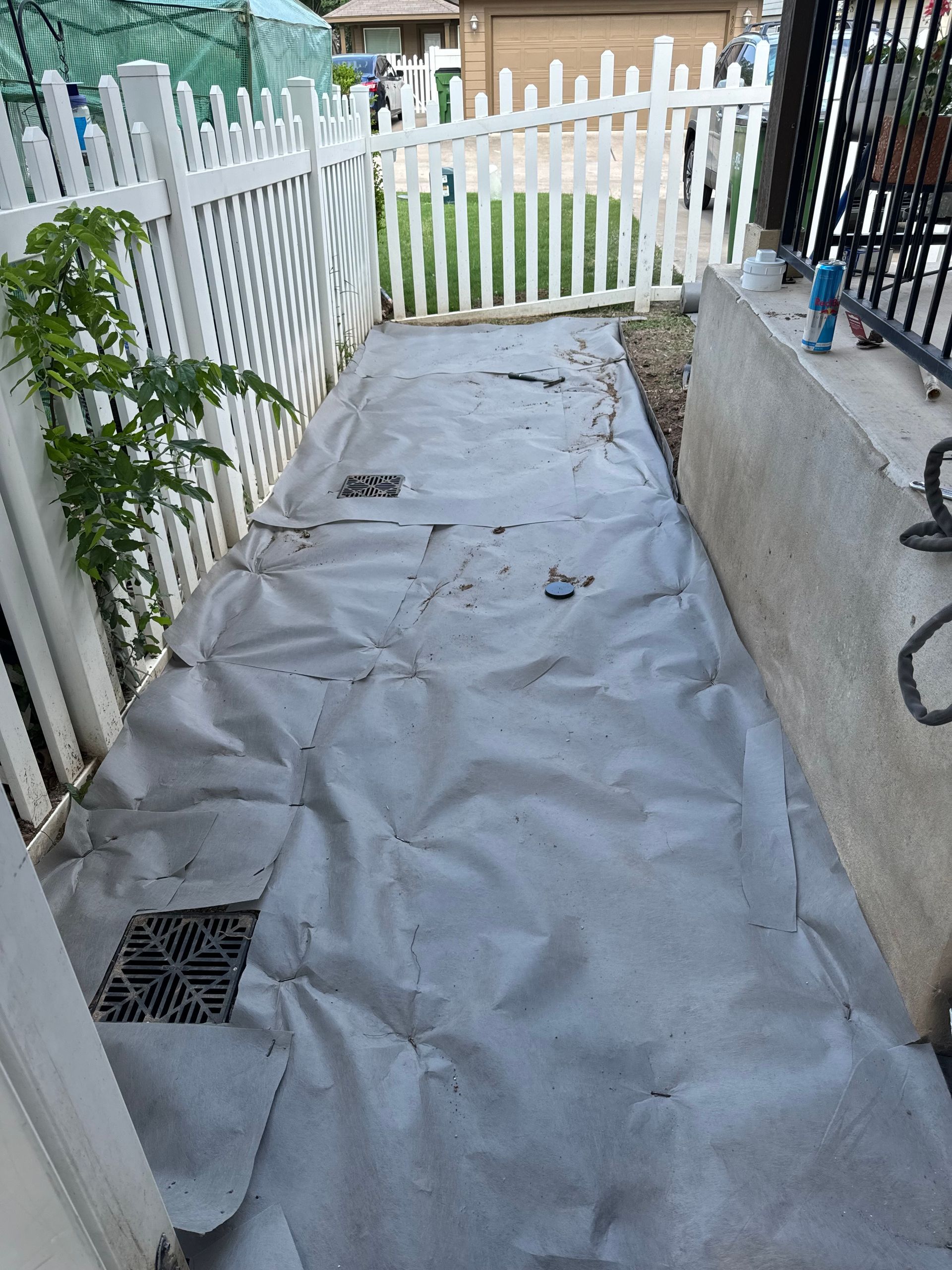 Narrow pathway with gray fabric ground cover, white picket fence, and a young plant.