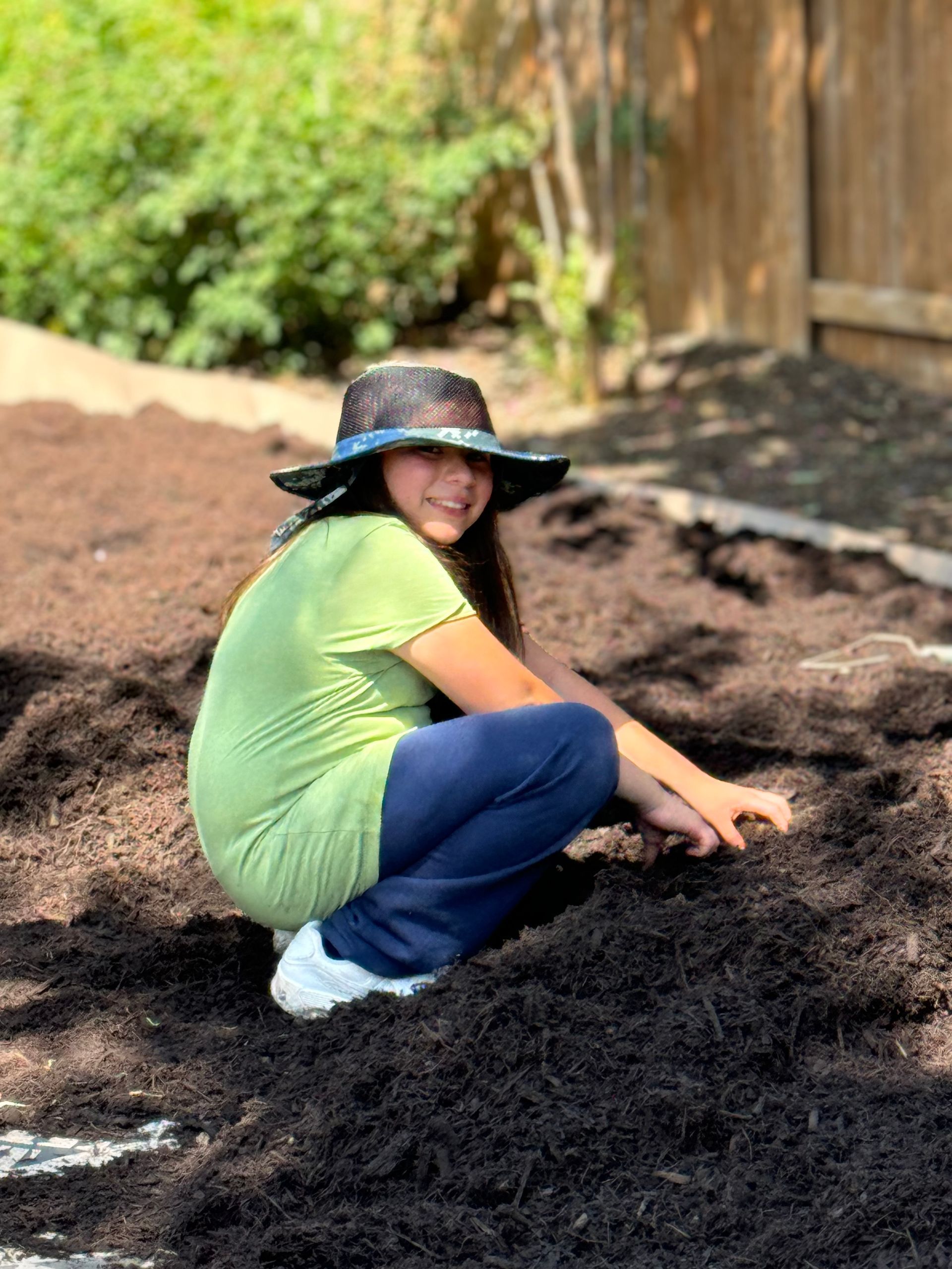 Person crouched in mulch, wearing a hat and green shirt, smiling 