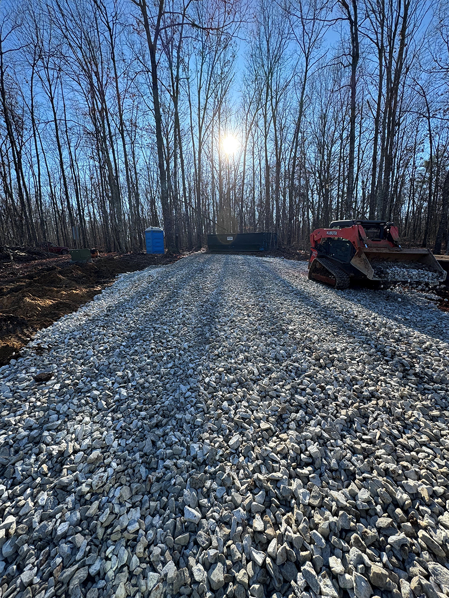 A gravel road with trees in the background and the sun shining through the trees.