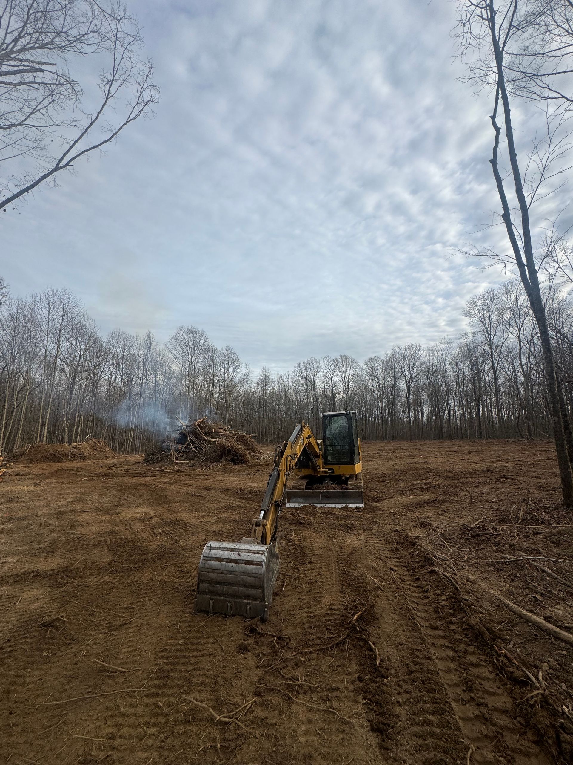 Excavator on a cleared land, smoke rising from a pile of debris, trees in the background under a cloudy sky.