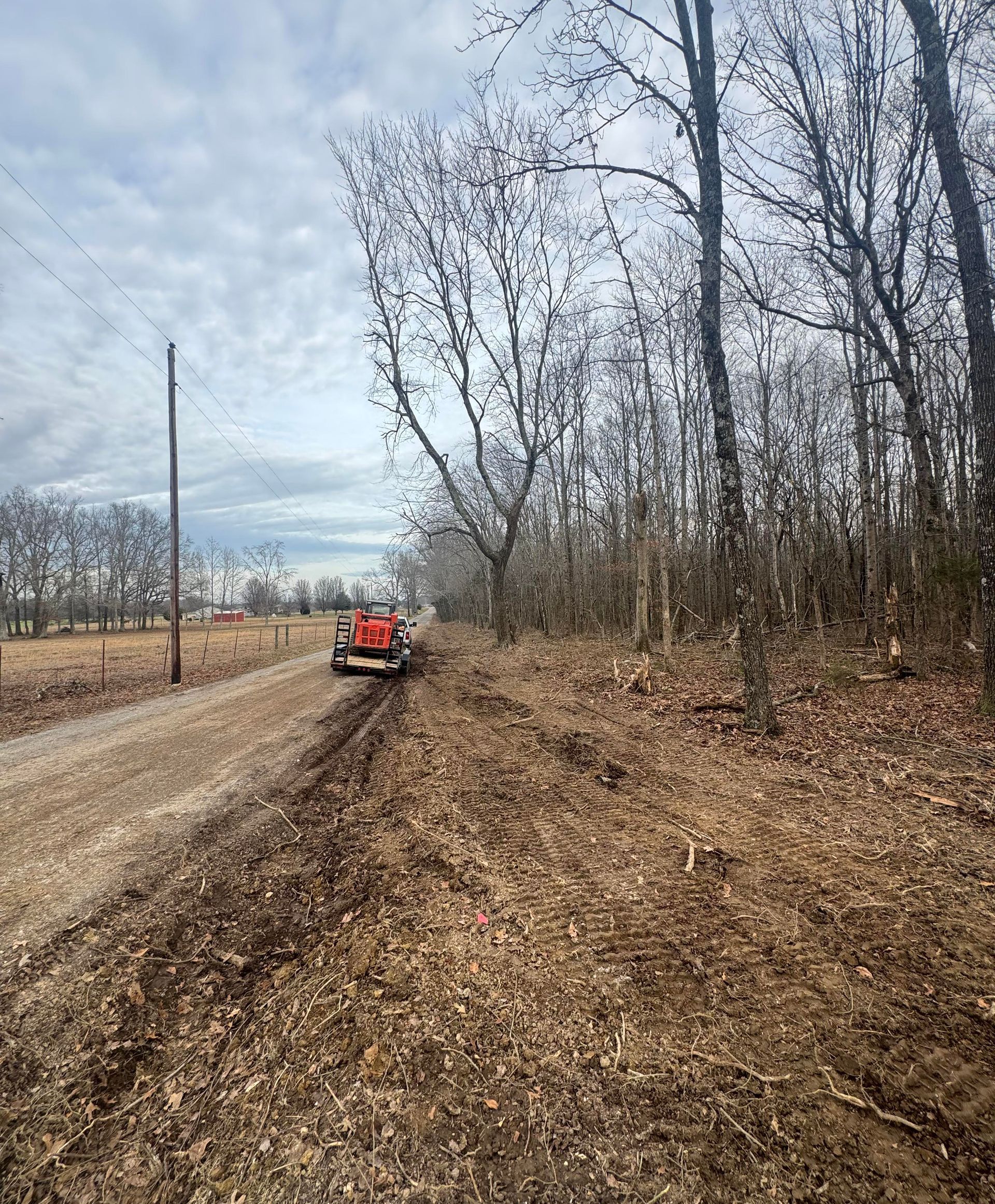 Dirt road with tractor clearing brush next to a wooded area under a cloudy sky.