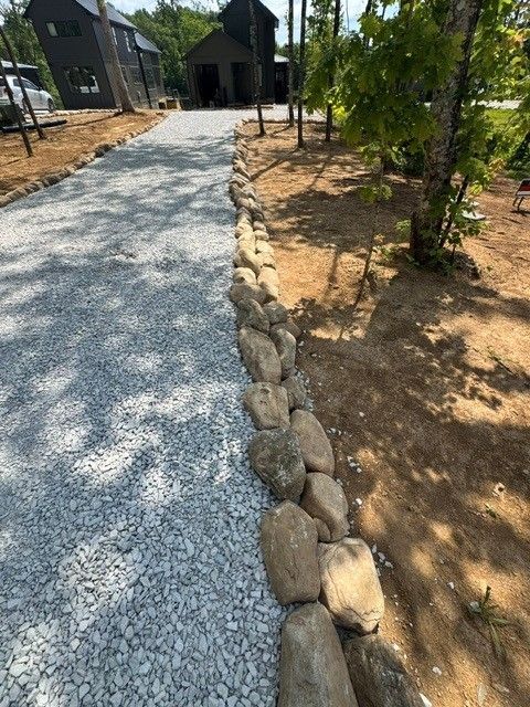 Gravel walkway bordered by rocks beside sandy landscaping and trees leading to a dark building entrance.