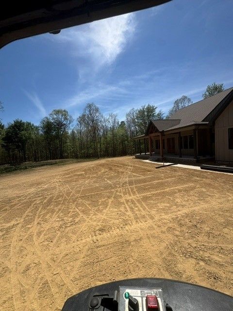 Graded dirt lot beside a house under construction, with trees and a blue sky in the background.