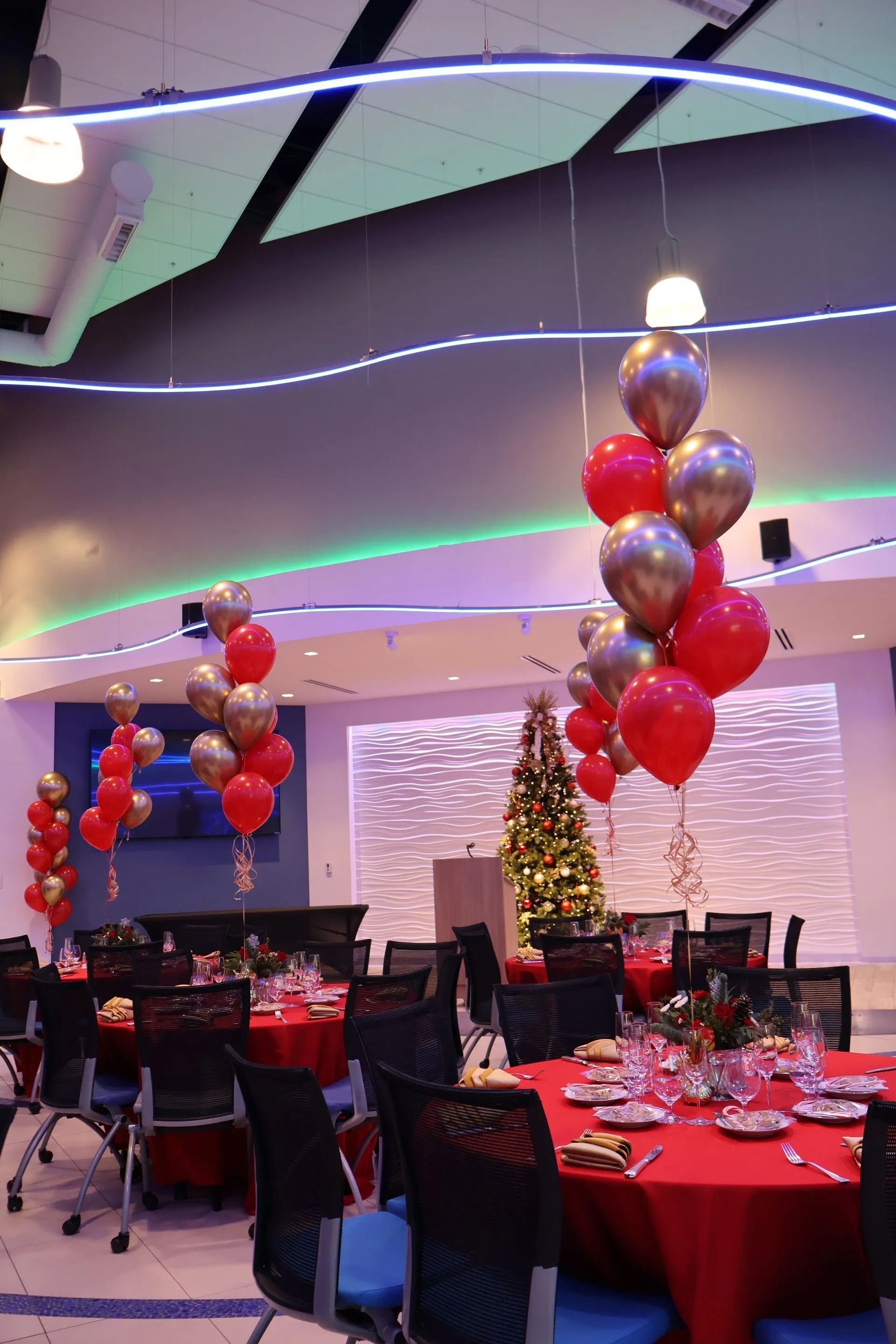 Decorated banquet hall with red table settings, balloon centerpieces, and a lit Christmas tree