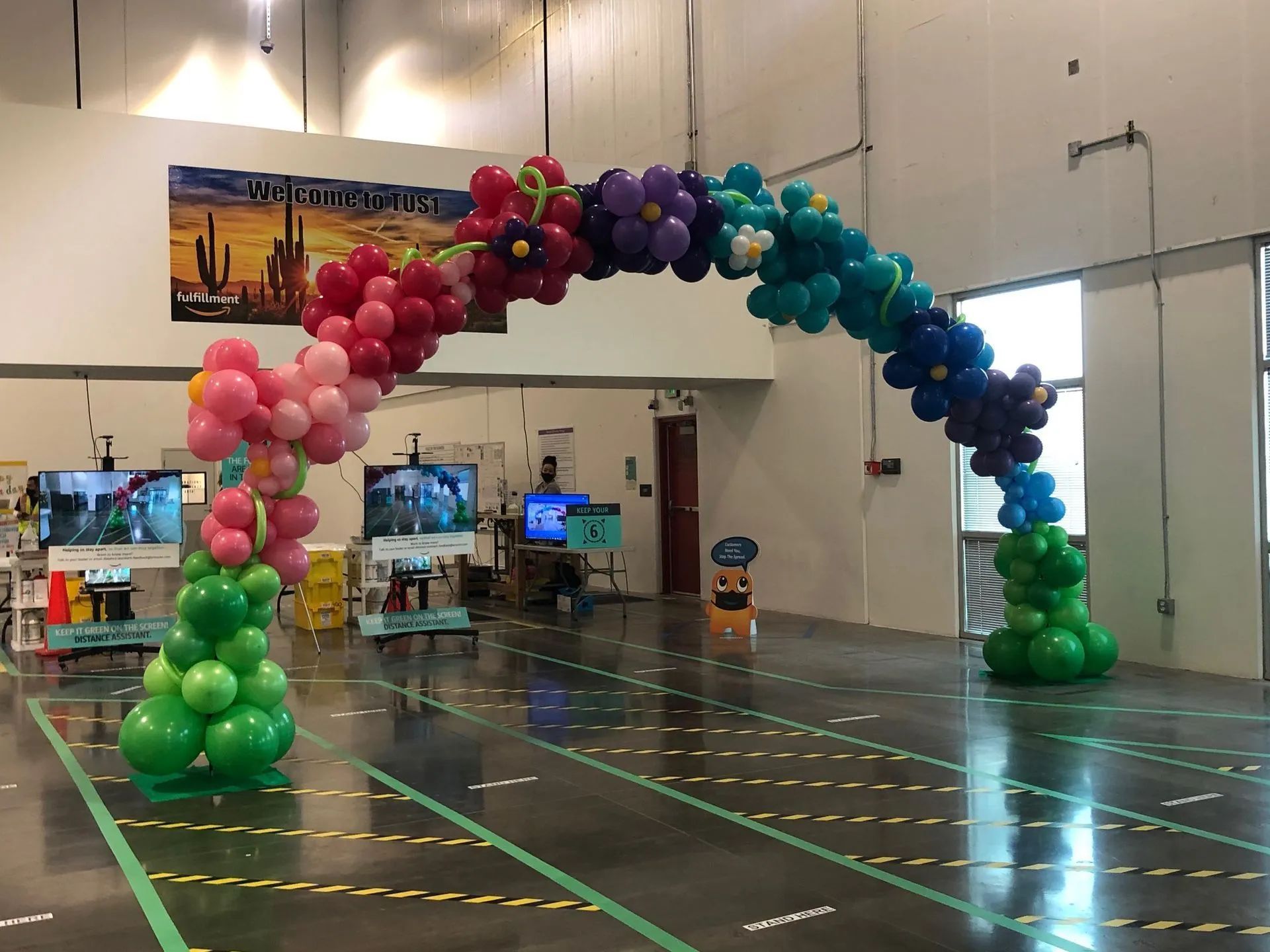 Colorful balloon arch over an indoor floor, with pink, green, purple, and blue balloons in a large hall.