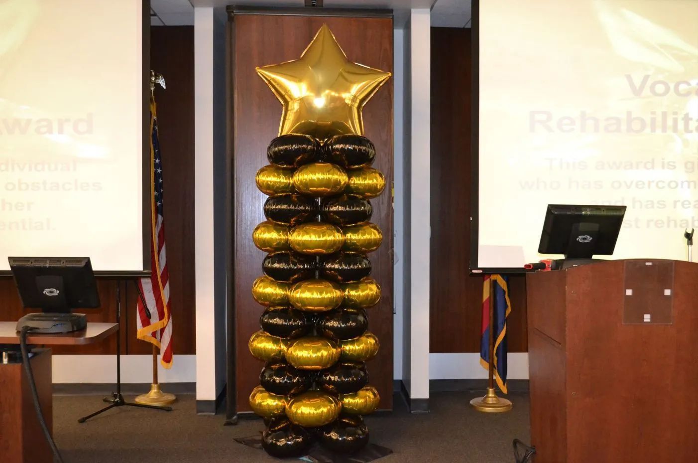 Gold star atop a stack of black and gold balloons on a stage with podiums and flags