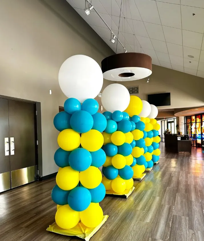 Blue, yellow, and white balloon columns in a hallway with wood floors and ceiling lights.