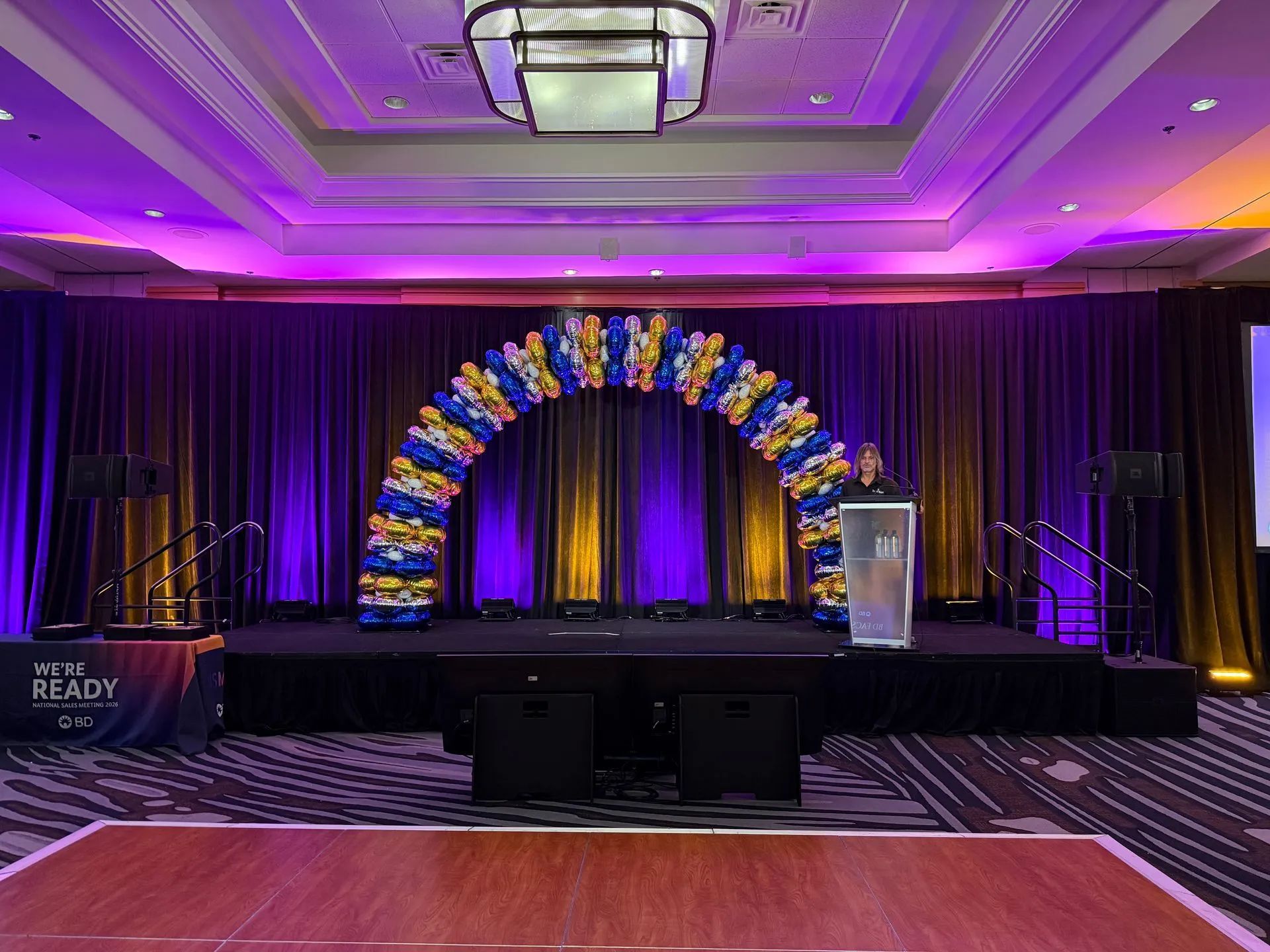 Purple-lit banquet stage with balloon arch, podium, and draped backdrop in a conference hall