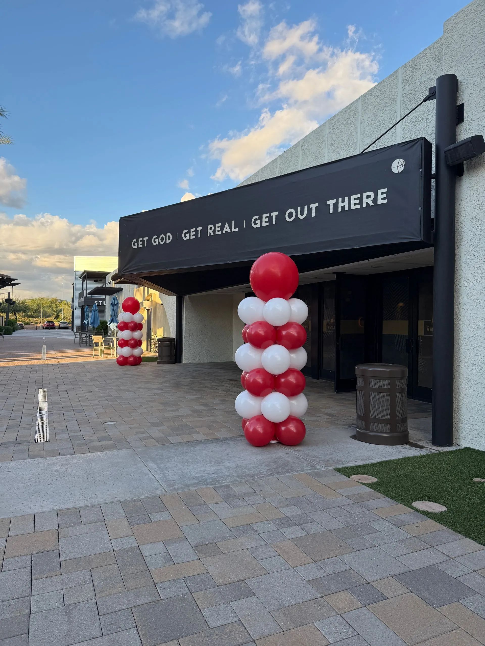 Outdoor building entrance with “Get out there” sign and red-and-white balloon columns at dusk.