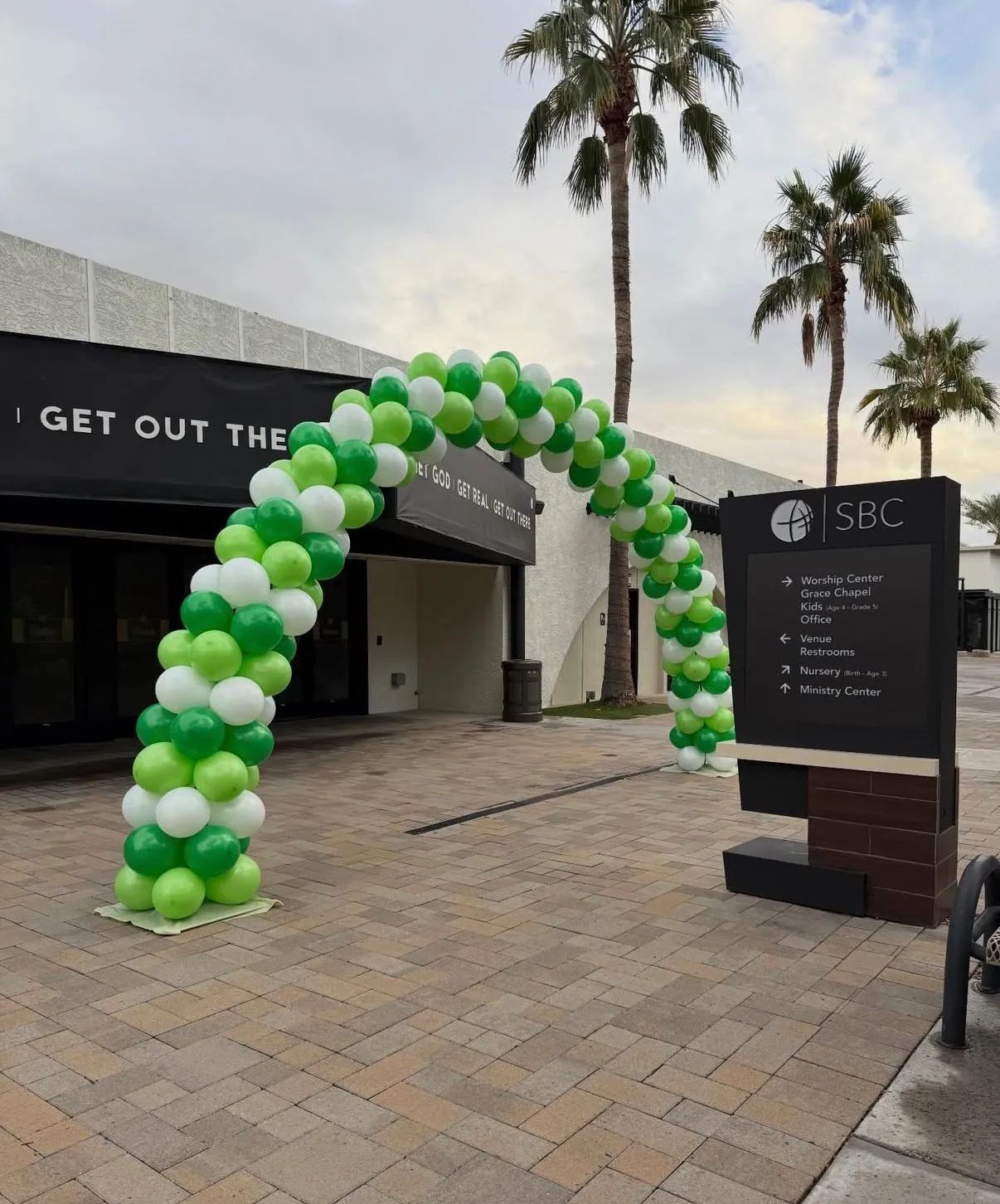 Green-and-white balloon arch outside a building entrance beside a black signboard and palm trees.