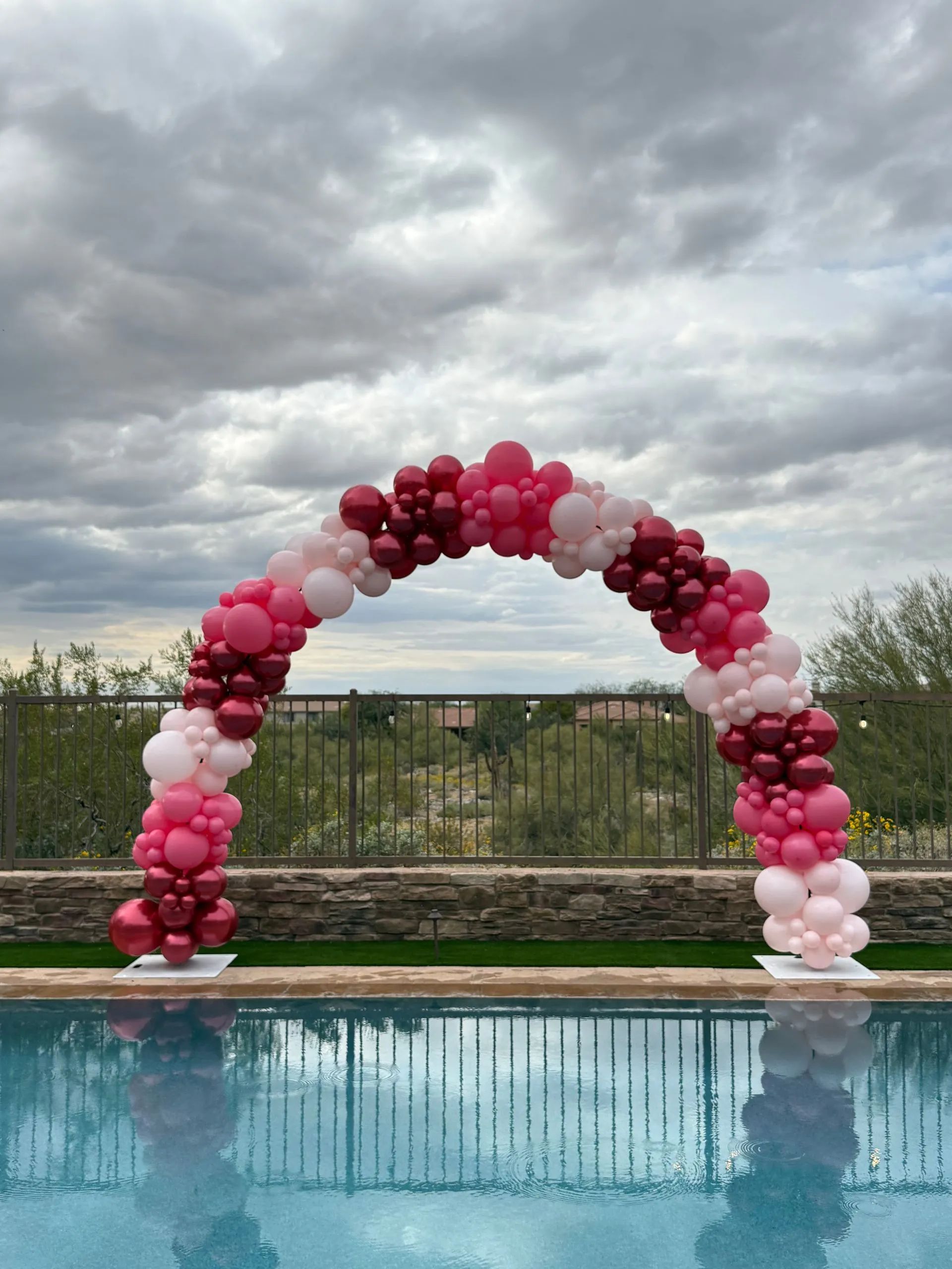 Pink and white balloon arch beside a pool under a cloudy sky