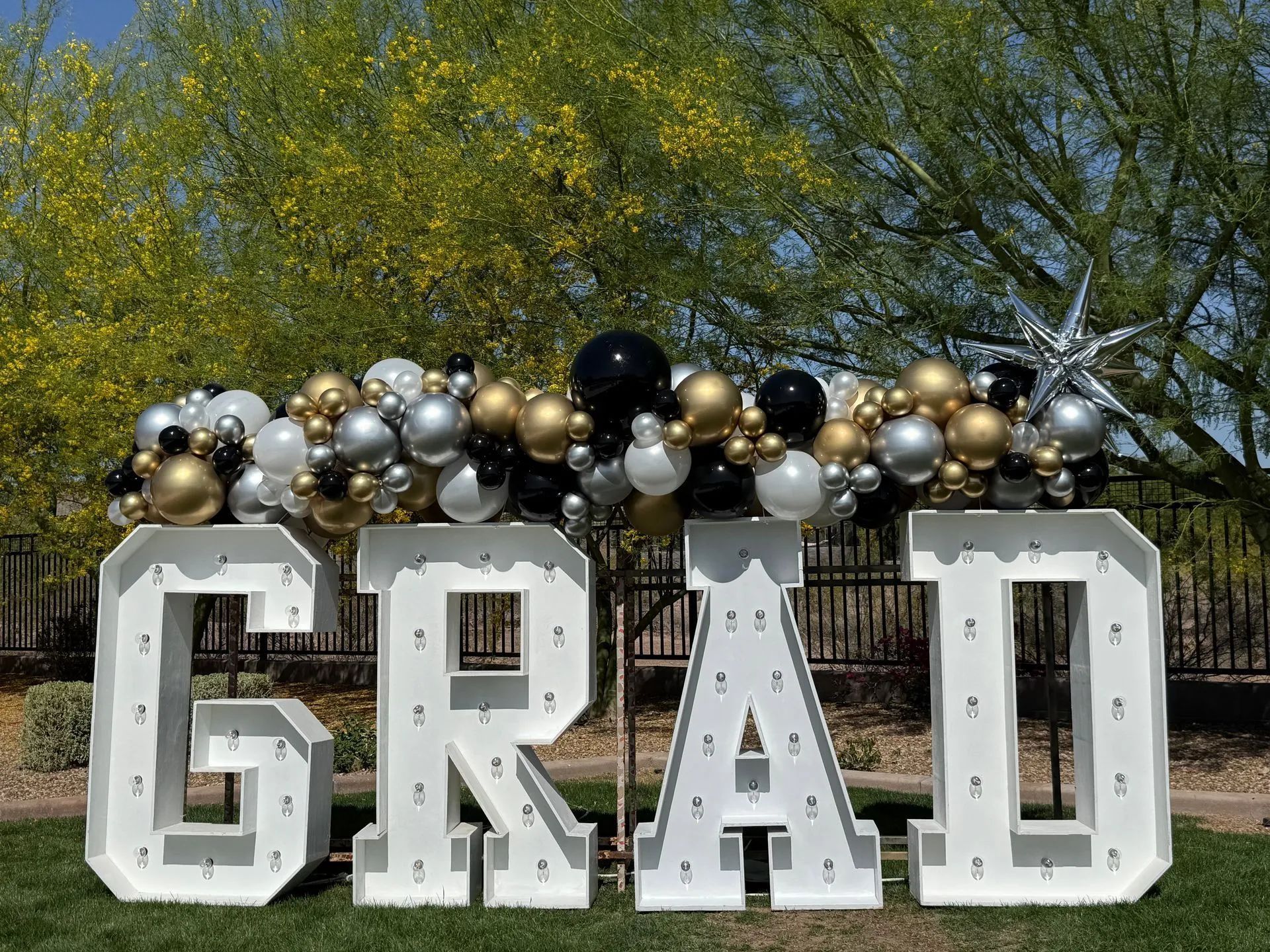 Large white “GRAD” letters decorated with black, gold, and silver balloons outdoors under trees.