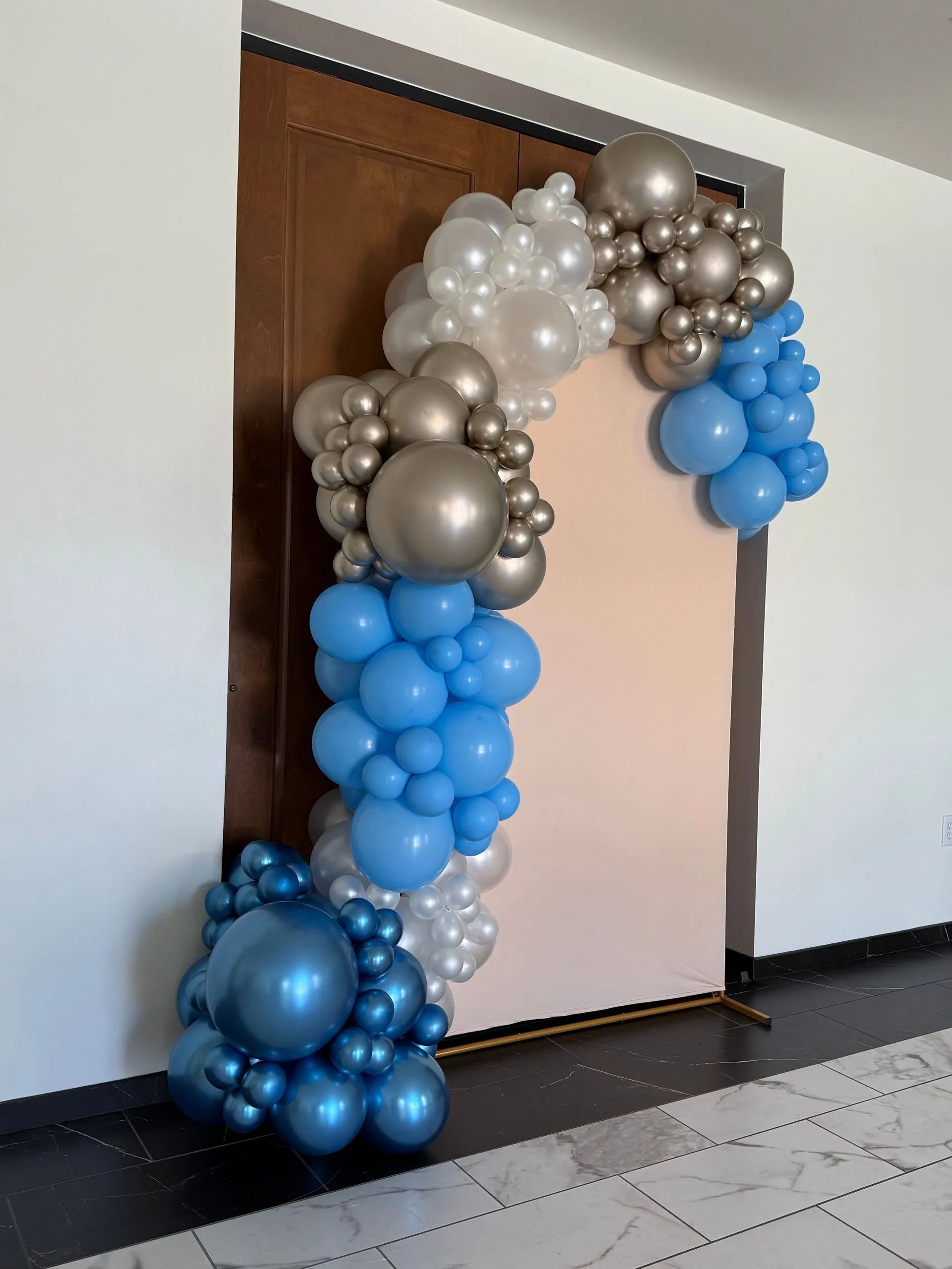 Balloon arch in blue, silver, and white framing a doorway in an indoor hallway.