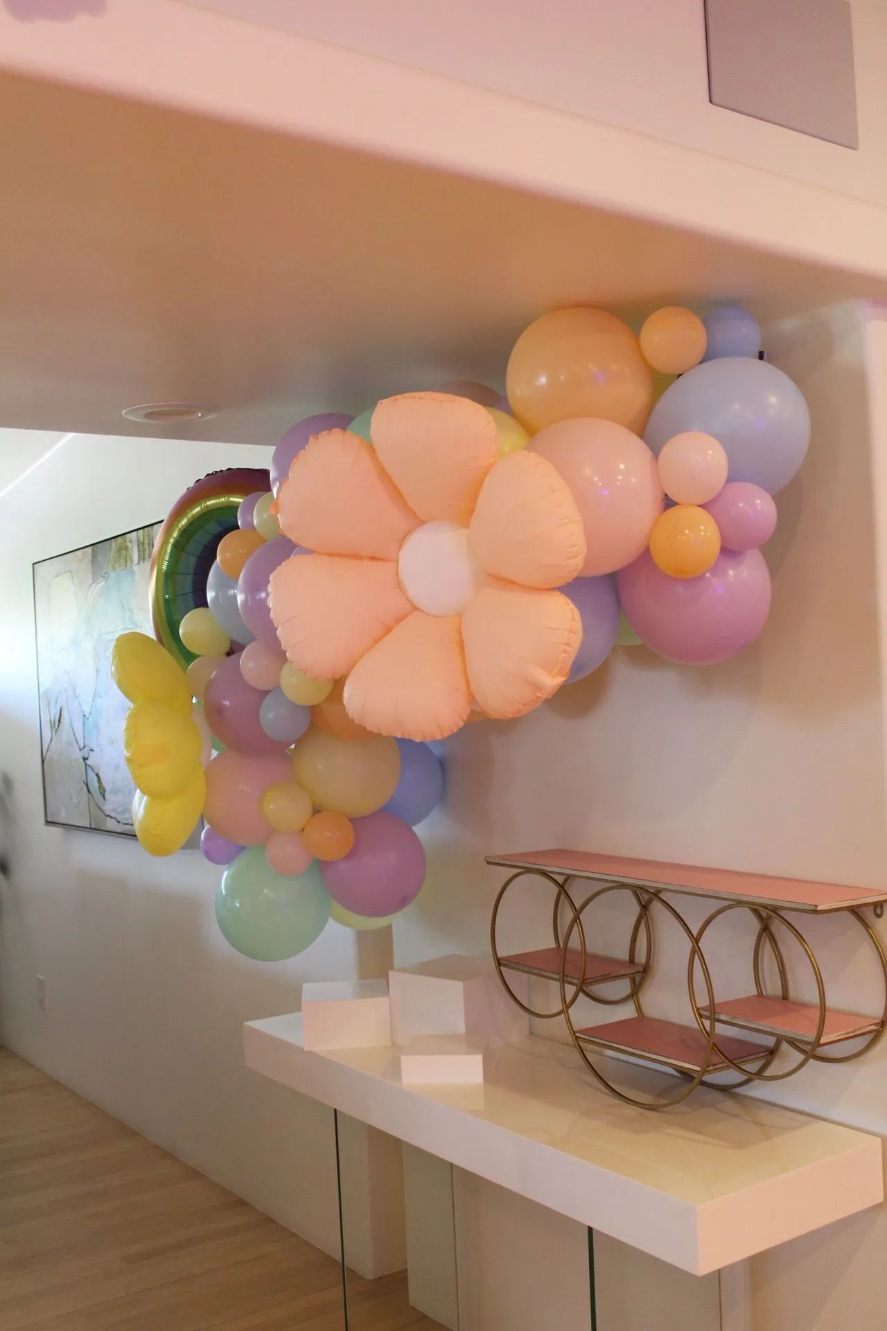 Pastel balloon flower installation in a hallway above white tables, with a rainbow balloon arch in the background