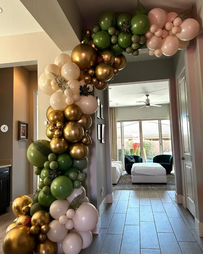 Balloon garland in green, gold, and blush drapes a hallway archway leading to a bright room.