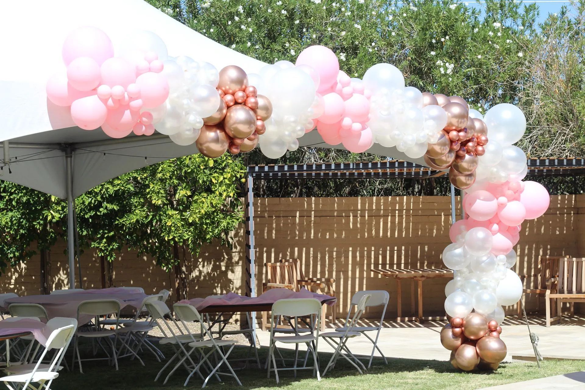 Outdoor event setup with pink, white, and gold balloon arch over tables and chairs in a shaded yard