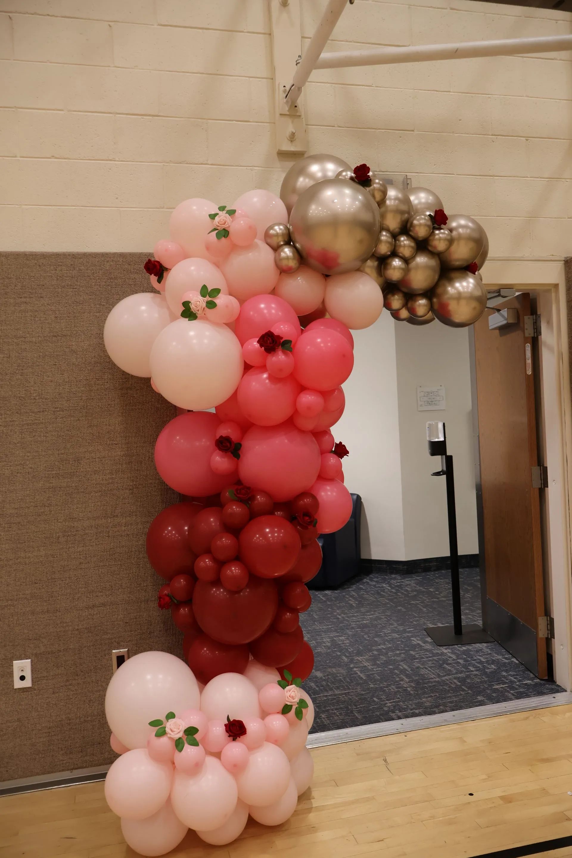 Pink, red, and gold balloon arch framing a doorway in an indoor event space