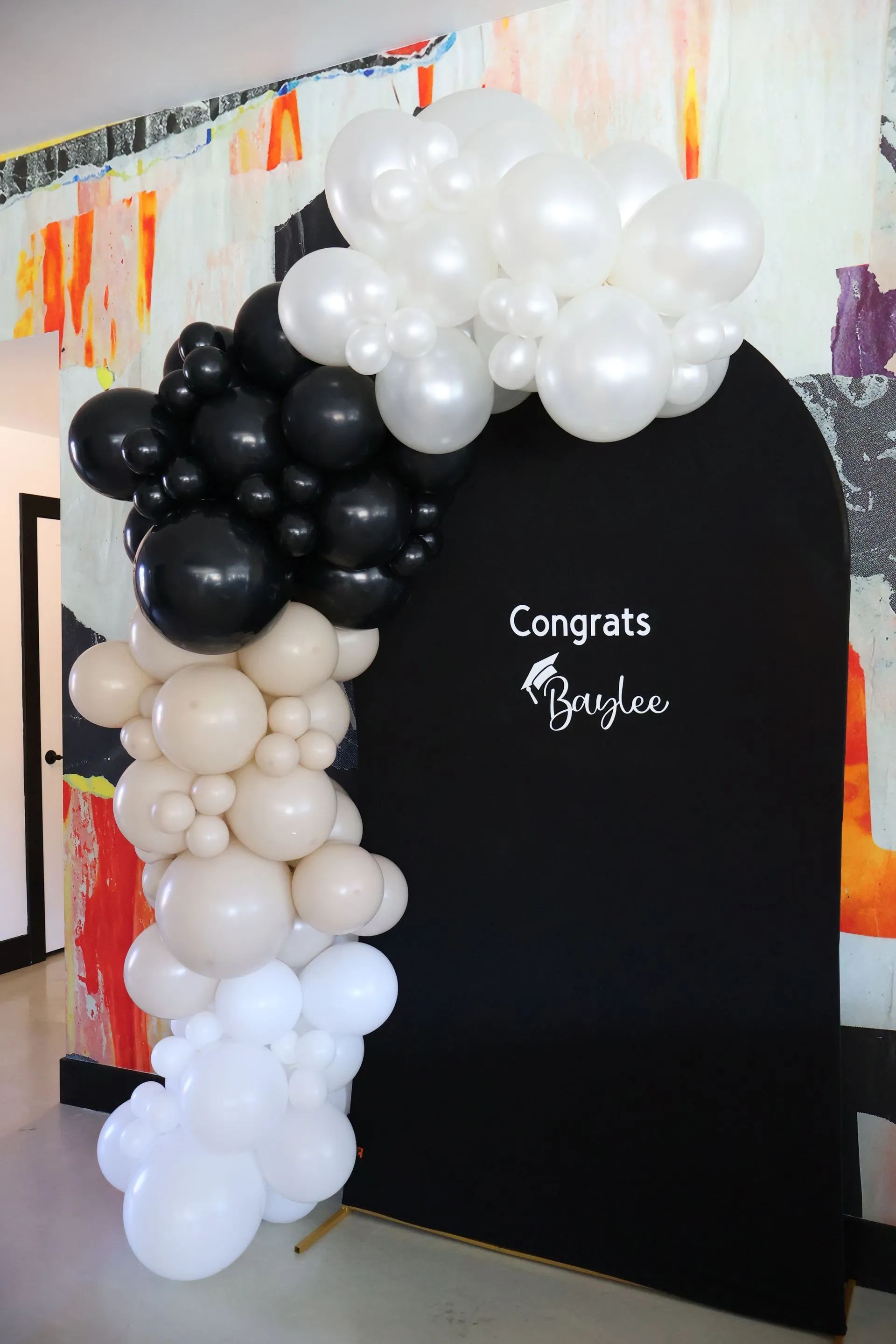 Black, white, and beige balloon arch around a black backdrop reading “Congrats” at an indoor event