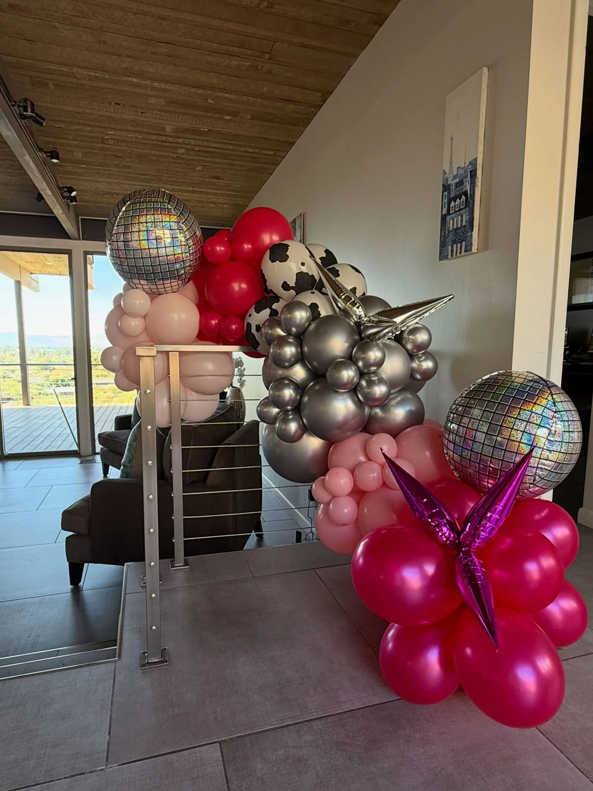 Colorful balloon arrangements lining a patio walkway, with silver, pink, red, and disco-ball balloons