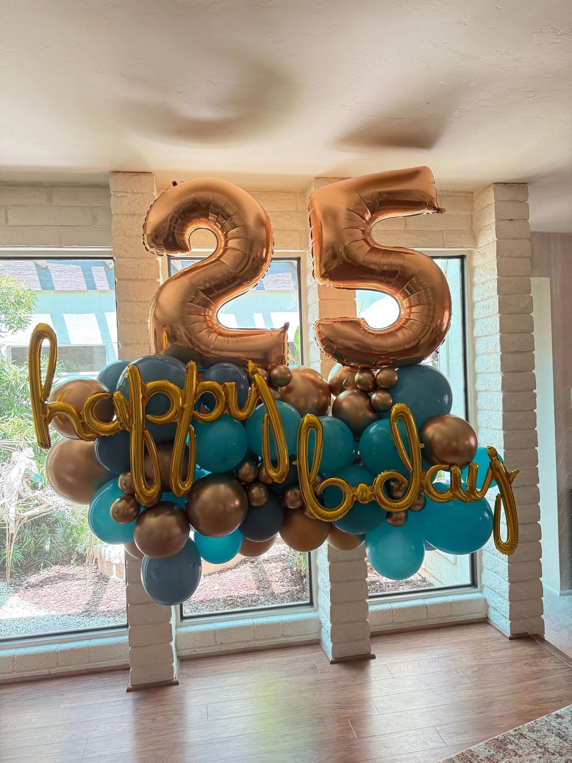 Gold and teal 25th birthday balloon display in a bright room with floor-to-ceiling windows