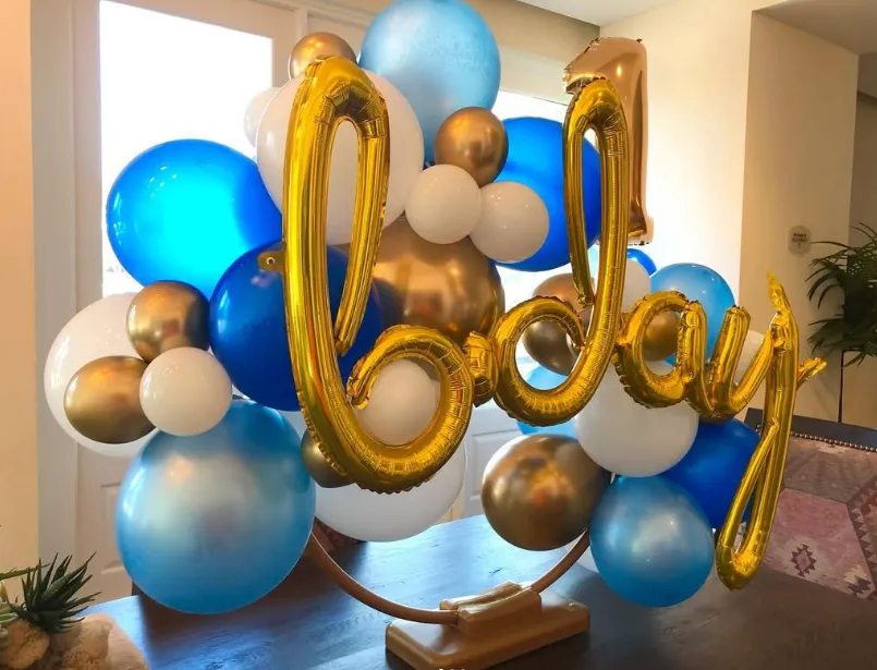 Gold cursive “baby” balloon display with blue, white, and gold balloons indoors